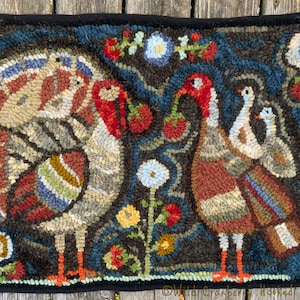 Op de afbeelding: Handgehaakte vloerkleed met zwarte rand, met twee kalkoenen en twee kuikens. De kalkoenen hebben rode koppen en kleurrijke staartveren. Het vloerkleed heeft bloemmotieven in rood, wit en geel, tegen een donkerblauwe en bruine achtergrond.