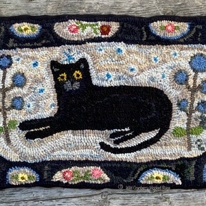 May include: A black cat with yellow eyes is lying down on a cream-coloured rug with blue flowers and a brown stem. The rug is surrounded by a black border with a floral design. The text "Wild Cranberry Hooked Rugs" is in the bottom right corner.