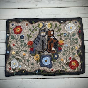 May include: Hand-hooked rug with a design of two cats and floral patterns. The cats are grey and brown, sitting together. The rug has a black border and a beige background with colourful flowers in red, blue, and yellow.