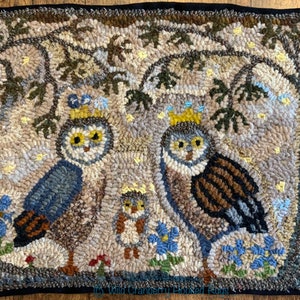 Op de afbeelding: Handgehaakte vloerkleed met een familie uilen onder een boom. De uilen hebben gele ogen en dragen kronen. Het vloerkleed heeft een neutraal kleurenpalet met blauwe en groene accenten. Het vloerkleed is rechthoekig en heeft een zwarte rand.