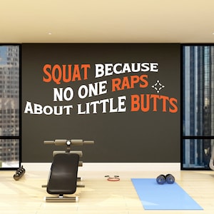 Può includere: Interno di palestra con tapis roulant, panca e manubri. Un tappetino da yoga blu e un muro con la scritta "SQUAT BECAUSE NO ONE RAPS ABOUT LITTLE BUTTS" in arancione e bianco. Grandi finestre offrono una vista sulla città.