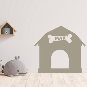May include: A grey dog house wall decal with a bone on top that says "MAX" in white letters. The decal is on a white wall with a wooden shelf above it.