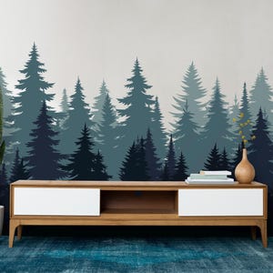 Könnte beinhalten: Ein Wohnzimmer-Interieur mit einem dunkelblauen und türkisfarbenen Wandaufkleber mit einer Waldsilhouette. Der Aufkleber zeigt verschiedene Kiefern in unterschiedlichen Größen. Vor der Wand steht ein Fernsehtisch aus Holz mit einer weißen Schublade. Links vom Tisch steht eine Topfpflanze mit einer Schlangenpflanze. Rechts vom Tisch steht eine Stehlampe aus Holz mit einem weißen Lampenschirm.
