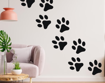May include: A white wall with a series of black paw prints in a diagonal line. The paw prints are of varying sizes and are arranged in a staggered pattern. There is a pink lamp hanging from the ceiling, a couch, a side table, and a shelf with a clock and other decorative items.