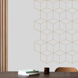 May include: A light grey wall with a gold geometric pattern of cubes. A wooden table with a stack of books, a candle holder, and a vase with a dried branch sits in front of the wall.