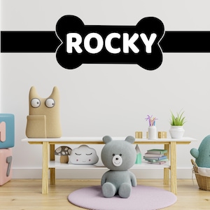 May include: A white wall with a black bone-shaped decal that says "ROCKY" in white letters. There is a white shelf with a stuffed bear, a mushroom, and a cloud on it. There are also two blocks with letters on them.
