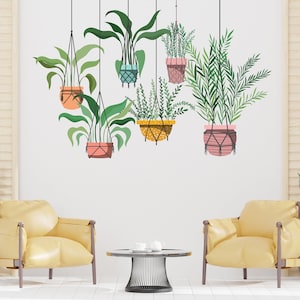 Op de afbeelding: Een witte muur met zeven hangende planten in macramé potten. De planten zijn in verschillende tinten groen en de potten zijn in tinten roze, oranje, geel en blauw. Twee gele fauteuils en een zwarte metalen tafel staan voor de muur.