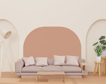 Puede incluir: Un sofá marrón claro con cuatro almohadas se encuentra frente a una pared de color melocotón con dos puertas arqueadas. Una lámpara de pie blanca con una pantalla tejida se encuentra a la izquierda del sofá. Dos plantas en maceta y una mesa auxiliar de madera se encuentran a la derecha del sofá.