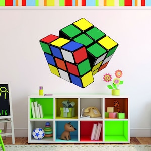 May include: A colorful 3D Rubik's Cube wall decal in a white room with a white shelf unit with colorful compartments. The shelf unit contains a variety of items, including books, a toy dog, a ball, and a phone.