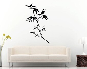 May include: Black silhouette of a bird perched on a bamboo branch with leaves. The artwork is mounted on a white wall above a white sofa. A vase with yellow flowers and a lamp are also visible.
