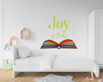 May include: A white wall with a green and rainbow-colored wall decal that says "Joy of Books" with a rainbow-colored open book with stars around it. A white bed with white bedding is in front of the wall.