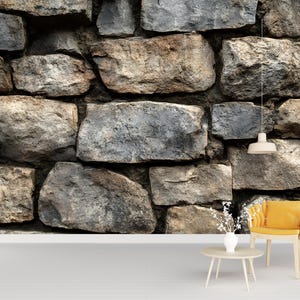 Op de afbeelding: Een stenen muur met verschillende tinten grijs en bruin, waardoor een textuur ontstaat. De muur is gemaakt van onregelmatig gevormde stenen, wat een rustieke en natuurlijke uitstraling geeft. Een kleine, ronde tafel, een gele stoel en een hanglamp staan voor de muur.