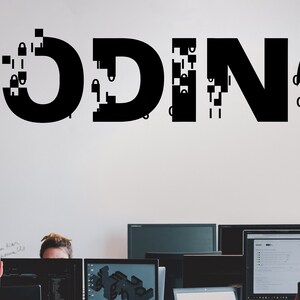 May include: The word "CODING" is written in black on a white wall, with some of the letters broken up into pixelated blocks.  Three people are sitting in front of computers, working on a project.