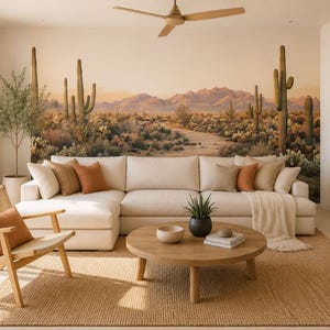 May include: A large wall mural depicting a desert landscape with saguaro cacti and mountains in warm beige and brown tones. The mural serves as a backdrop for a cream-colored sectional sofa, a round wooden coffee table, and a woven rattan chair.