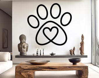 May include: A large black paw print wall decal with a heart in the center. The decal is on a white wall in a modern living room. The room also features a wooden coffee table, sculptures, and candles.
