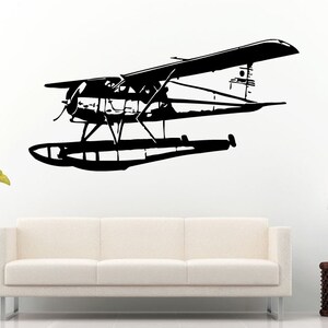 May include: Black and white silhouette of a seaplane with two pontoons. The seaplane is flying in the air. The image is on a white background.