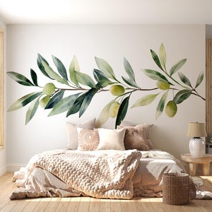 Può includere: Una camera da letto con un murale a forma di ramo d'ulivo con foglie e olive verdi. Il letto è fatto con una coperta beige lavorata a maglia e cuscini. Un comodino in legno con una lampada e un cesto di vimini completano l'arredamento.