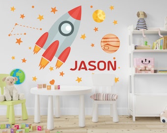 May include: A white wall with a rocket ship, planets, stars, and the name "JASON" in red letters. There is a green chair with a stuffed lamb on it, a white table with wooden blocks, and a white bookshelf with books and a pink storage box.