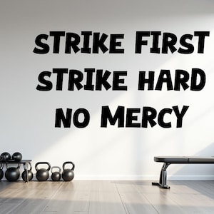 May include: A white wall in a gym setting with the motivational phrase "STRIKE FIRST STRIKE HARD NO MERCY" in bold black letters. The room includes exercise equipment like kettlebells and a weight bench.