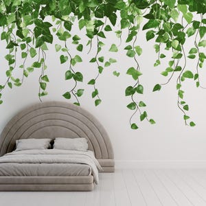 May include: A bedroom scene featuring a bed with a curved headboard, a large potted plant, and hanging green vines. The bed has a gray comforter and pillows. The plant is in a white pot on a wooden stand. The vines are a vibrant green.