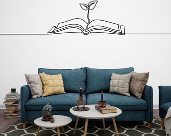 May include: A blue sofa with two throw pillows and a book with a plant growing out of it drawn on the wall above it. There are two coffee tables in front of the sofa, one with a glass cloche with pine cones inside and the other with a book and two candle holders.