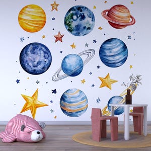 Può includere: Decorazioni murali in stile acquerello raffiguranti pianeti e stelle blu, gialle e rosse. Un tavolino bianco e due sedie rosa sono di fronte al muro. Un cuscino a forma di orso a righe rosa e bianche è sul pavimento.