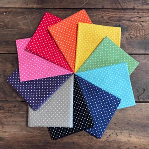May include: A collection of colorful fabric squares arranged in a circular pattern. The fabrics are in various colors, including red, orange, yellow, green, blue, and purple, each with a white polka dot pattern. The fabrics are displayed on a wooden surface.
