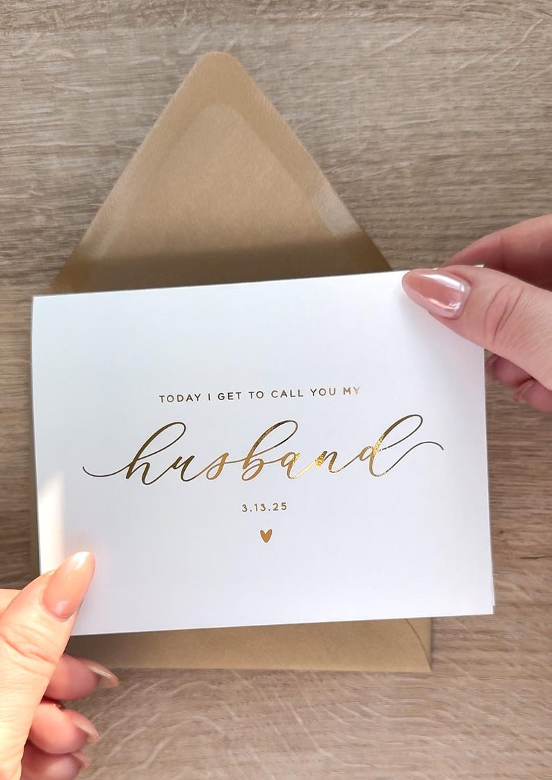 Personalized wedding day card reading “Today I Get to Call You My Husband” in gold foil with custom date, modern minimalist bride to groom wedding card held above kraft envelope.