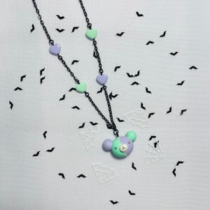 May include: A black chain necklace with a green and purple bear charm. The necklace has several pastel green and purple heart charms.