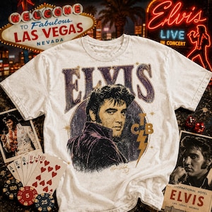 May include: A white short-sleeve t-shirt featuring a graphic of Elvis Presley in a dark purple jacket. The text ELVIS appears in large purple letters above the portrait, with the letters TCB and a lightning bolt symbol in gold to the right.