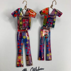 May include: A pair of dangle earrings shaped like a shirt and pants set, featuring a glossy finish with a multi-colored abstract pattern of red, yellow, and blue geometric shapes and dots. The earrings hang from gold-toned metal hooks.