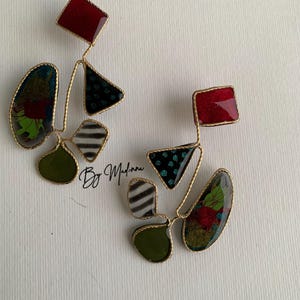 May include: A pair of dangle earrings featuring multiple geometric shapes outlined in gold wire. Each earring includes a red square, a black triangle with teal dots, a black and white striped shape, and an olive green oval, plus a floral pattern.