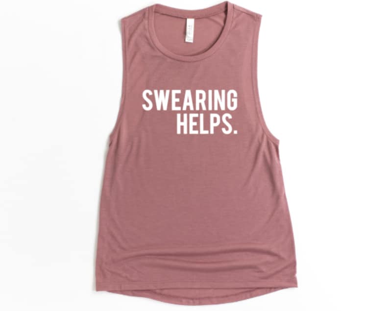 May include: A sleeveless, dusty rose-colored tank top with the white text "SWEARING HELPS." The shirt has a scoop neck and a relaxed fit. This casual top is suitable for workouts or everyday wear.
