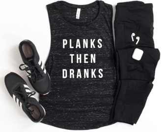 May include: Black tank top with the text "Planks Then Dranks" in white. The tank top is on a white background with black sneakers and black leggings.