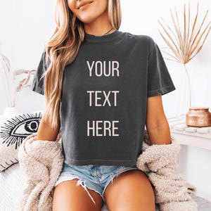 K&ouml;nnte beinhalten: Dunkelgraues T-Shirt mit den Worten "YOUR TEXT HERE" in wei&szlig;en Blockbuchstaben. Das Shirt ist kurz&auml;rmlig und hat einen Rundhalsausschnitt. Das Model tr&auml;gt Jeansshorts und eine cremefarbene Strickjacke.