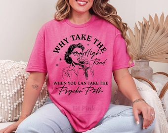 May include: A bright pink t-shirt with the text "WHY TAKE THE High Road WHEN YOU CAN TAKE THE Psycho Path" in black. The shirt features a black and white illustration of a woman wearing glasses and drinking from a glass. The shirt is a comfortable fit.