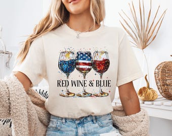 May include: A cream-colored t-shirt featuring a graphic of three wine glasses filled with red, white, and blue colors, including an American flag pattern in the center glass. The text "RED WINE & BLUE" is printed in black below the glasses.