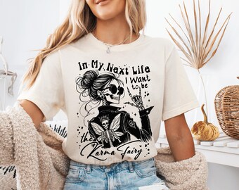 May include: Cream-colored t-shirt featuring a black graphic of a skeleton woman wearing sunglasses and a leather jacket. The text reads "In My Next Life I want to be the Karma Fairy."
