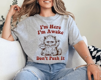 May include: A heather gray short-sleeve t-shirt featuring a brown graphic of a grumpy, fluffy cat holding a coffee mug. The text above the cat reads I'm Here I'm Awake and the text below the cat reads Don't Push It in a bold, dark red font.