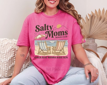 May include: A hot pink t-shirt with the text "Salty Moms Beach Club" above a beach scene with two beach chairs and an umbrella. The phrase "GIRLS JUST WANNA HAVE SUN" is printed below the beach scene.