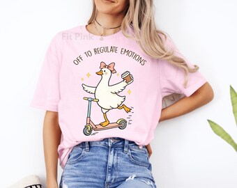 May include: Pink t-shirt with the text "OFF TO REGULATE EMOTIONS". The shirt features a cartoon goose riding a pink scooter while holding a coffee cup. The goose has a pink bow and is surrounded by yellow stars.