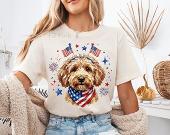 May include: Cream-colored t-shirt featuring a dog illustration with patriotic elements. The dog wears a headband and bandana with the American flag design. The design includes red, white, and blue stars, fireworks, and American flags.