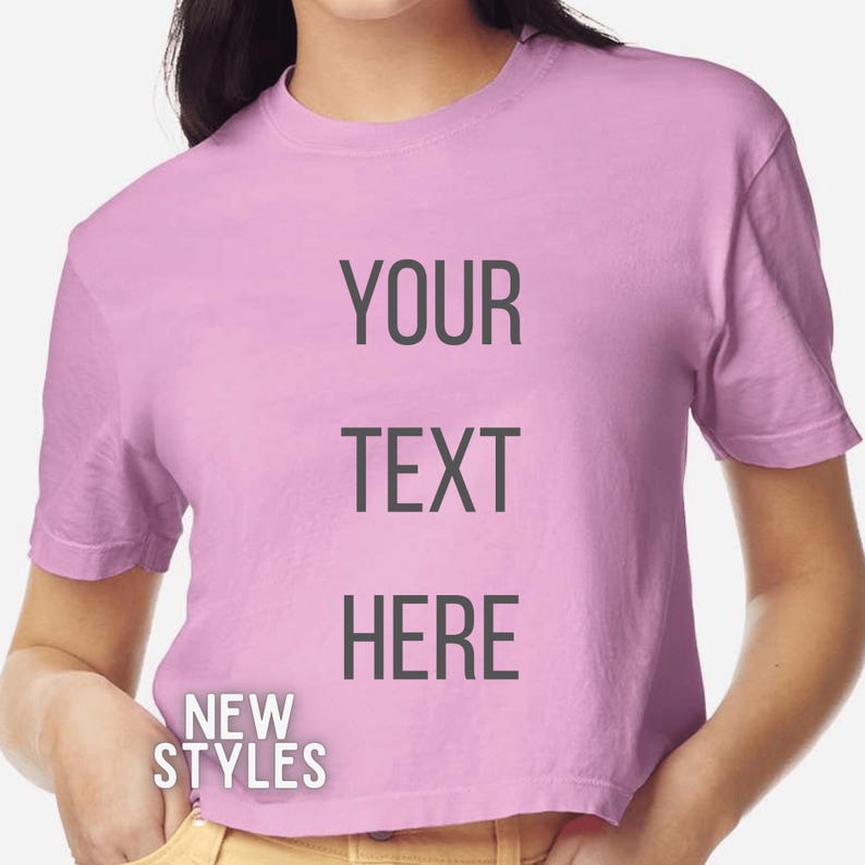K&ouml;nnte beinhalten: Ein hellrosa Crop-Top-T-Shirt mit dem Text "YOUR TEXT HERE" in Dunkelgrau. Die Worte "NEW STYLES" sind unten auf dem Shirt in Wei&szlig; gedruckt.