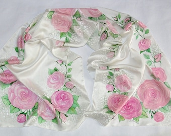 May include: A white silk scarf with a hand-painted design of pink roses and green leaves. The roses are arranged in a flowing pattern across the scarf.