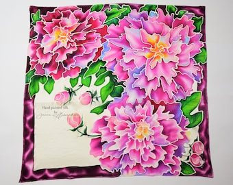 May include: Hand-painted silk scarf featuring a vibrant floral design with pink peonies and green leaves. The scarf has a white background with a burgundy border and the text "Hand painted silk by Janna Albrakhon" in the lower left corner.