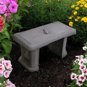 May include: A gray concrete garden bench with ornate detailing, including a decorative emblem on the top. The bench is surrounded by colorful flowers, including pink, yellow, and white blooms, creating a vibrant outdoor scene.