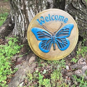 May include: A round, golden-colored garden stone with a blue butterfly design and the word "Welcome" in blue script. The stone is placed near a tree trunk and surrounded by green grass and foliage.
