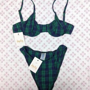 May include: A two-piece bikini set featuring a dark green and navy blue plaid pattern. The top has underwire cups and thin adjustable straps. The high-cut bottom includes a white tag with the brand name Jesilis printed in gold script.
