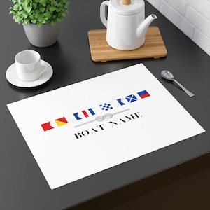 May include: White placemat with a nautical theme featuring a rope knot and a line of colorful signal flags. The text "BOAT NAME" is printed below the knot.