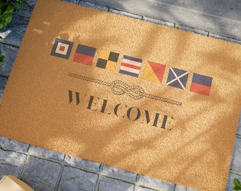 May include: A brown coir doormat with a welcome message and a row of colorful nautical flags. The flags are red, white, blue, yellow, and black.